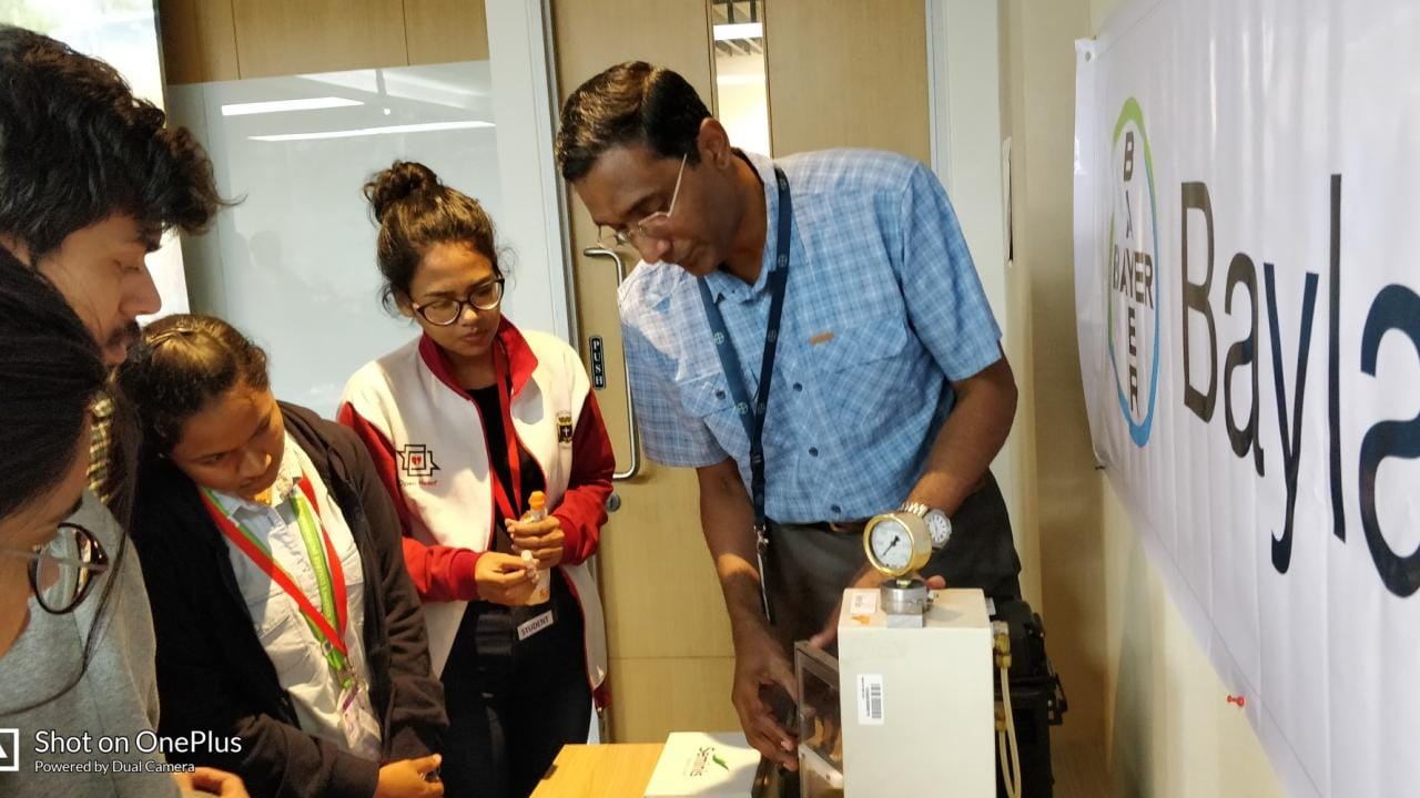 A group of people watches a man demonstrating a device in a room with a banner labeled "Dayla.