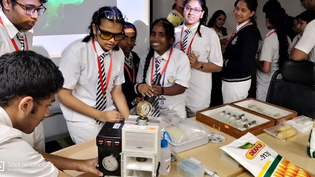 Students in school uniforms gather around a lab table with scientific equipment and materials, including a microscope and a gold detector. A teacher observes from the background.