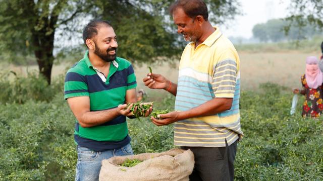 Two men smiling and holding green chili peppers in a field, with a sack of peppers in front of them.