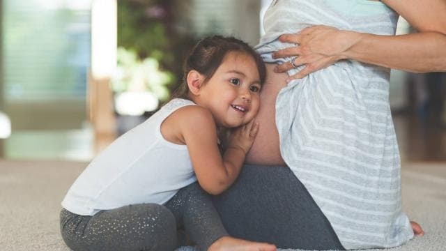 A young girl smiles and rests her head on a pregnant woman's belly, who is sitting on the floor.