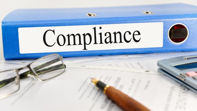 A blue binder labeled "Compliance" lies on a table next to glasses, documents, and a brown pen.