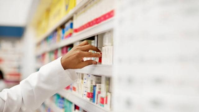 A pharmacist's hand reaches for a box on a pharmacy shelf filled with various medication packages.