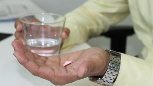 A person in a yellow shirt holding a pink pill in one hand and a glass of water in the other.