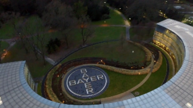 Aerial view of a circular Bayer logo on a grass area, surrounded by modern curved glass buildings, with trees and pathways in the background.