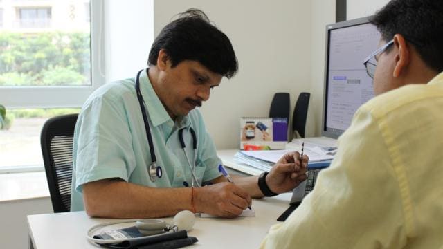 A doctor wearing a stethoscope sits at a desk, writing notes while talking to a patient.