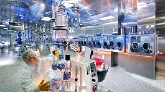 A scientist in protective gear works with complex glass apparatus in a high-tech laboratory. The room is equipped with advanced machinery and stainless steel surfaces.
