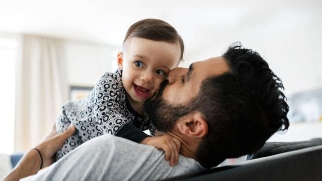 A man with a beard kisses a smiling baby wearing a patterned outfit while lying on a couch.