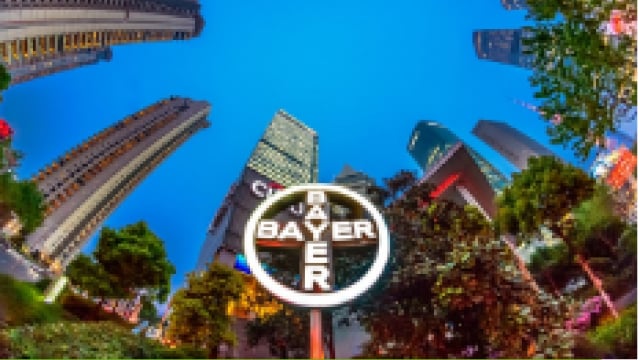 Fisheye view of skyscrapers and a large Bayer logo sign in an urban setting with trees at the bottom.