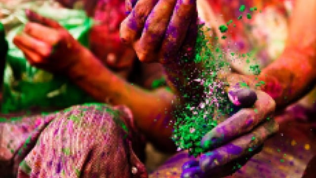 Hands covered in vibrant colored powders, one hand holding a mix of green powder, against a blurred background of colorful surroundings.