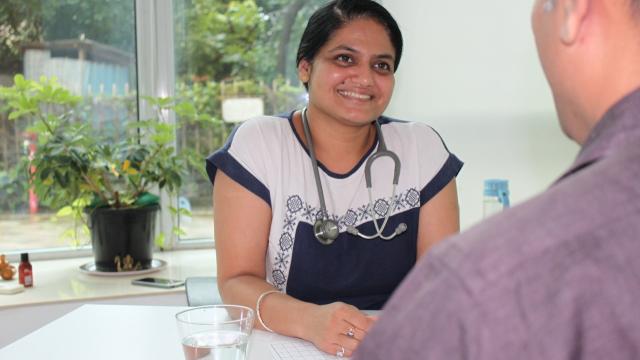 A doctor with a stethoscope smiles while talking to a patient in an office setting, with a glass of water and a plant nearby.