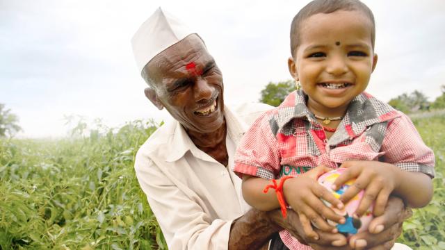 An elderly man wearing a white cap holds a smiling child in a field. The child holds a colorful ball. They both appear joyful against the backdrop of greenery.
