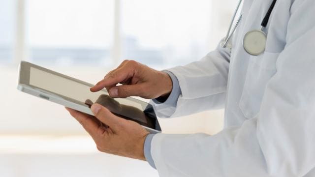A doctor in a white coat uses a tablet, with a stethoscope around their neck.