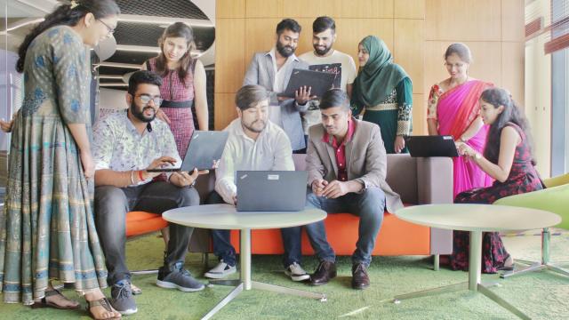 A group of people gathered in an office lounge, engaged with laptops and papers, some seated and others standing.