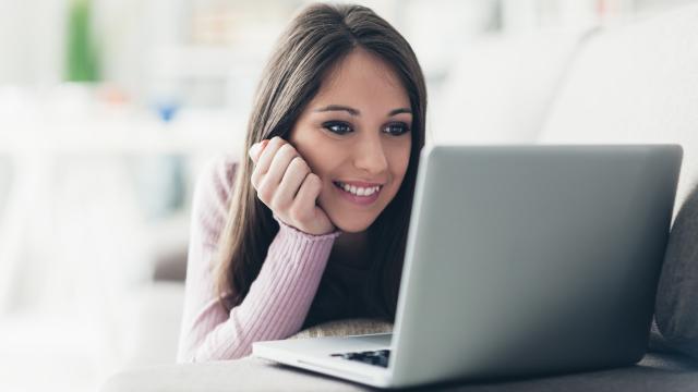 A woman with long brown hair smiles while looking at a laptop screen in a bright room.