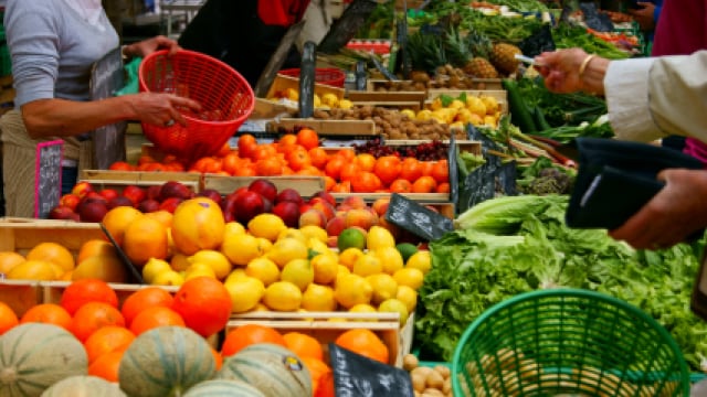A vibrant market stall displays a variety of fruits and vegetables, including oranges, lemons, apples, lettuce, and melons. People are selecting produce from baskets.