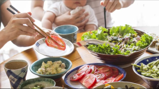 Hands using chopsticks to eat a meal with salad, sliced tomatoes, and various dishes on a table. A baby is held by someone in the background.
