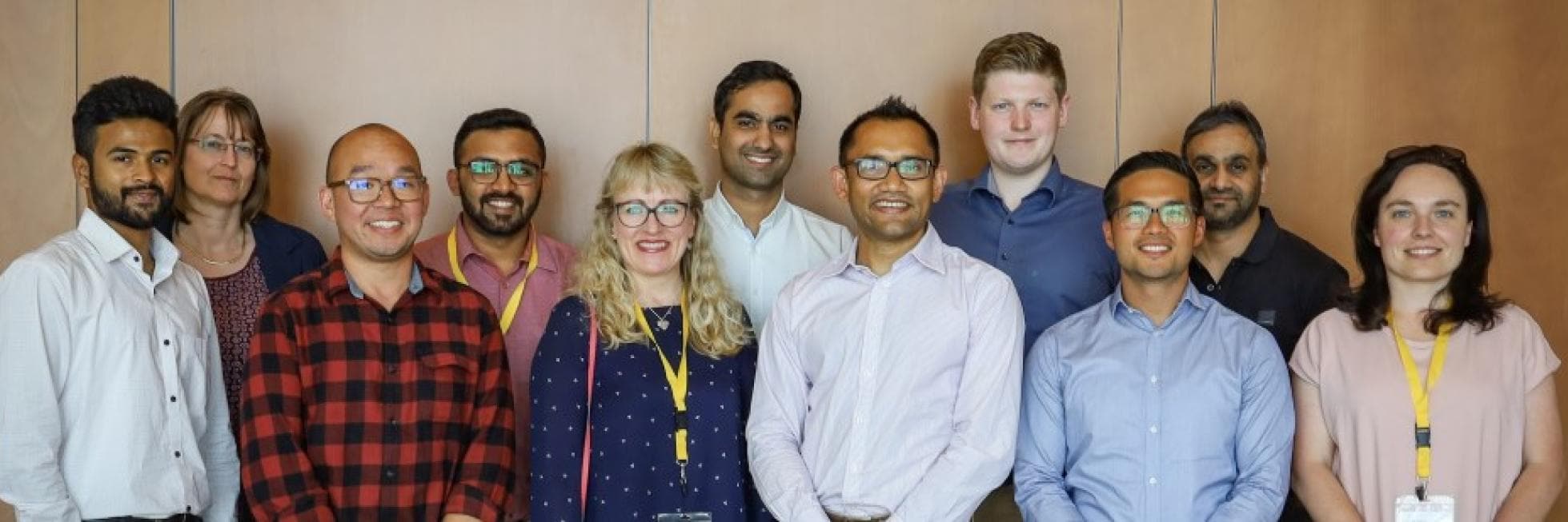 A group of twelve people of diverse backgrounds pose together in front of a wooden wall, smiling for the camera.
