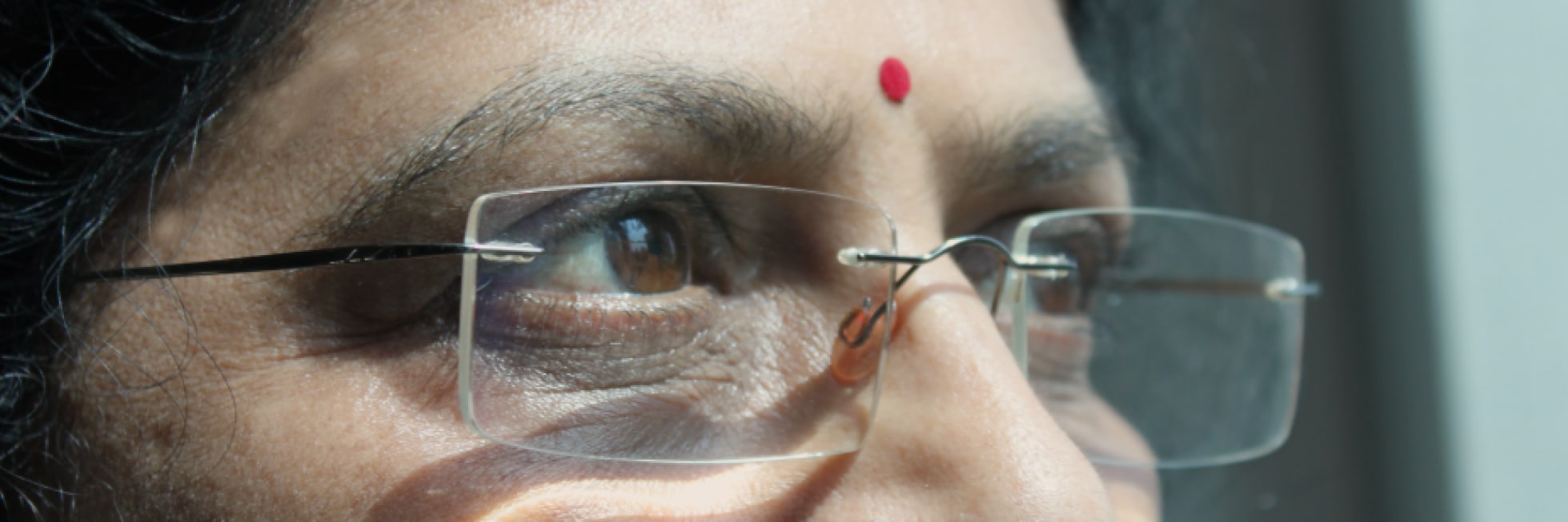 Close-up view of a person wearing glasses, looking out a window. A red bindi is on their forehead.
