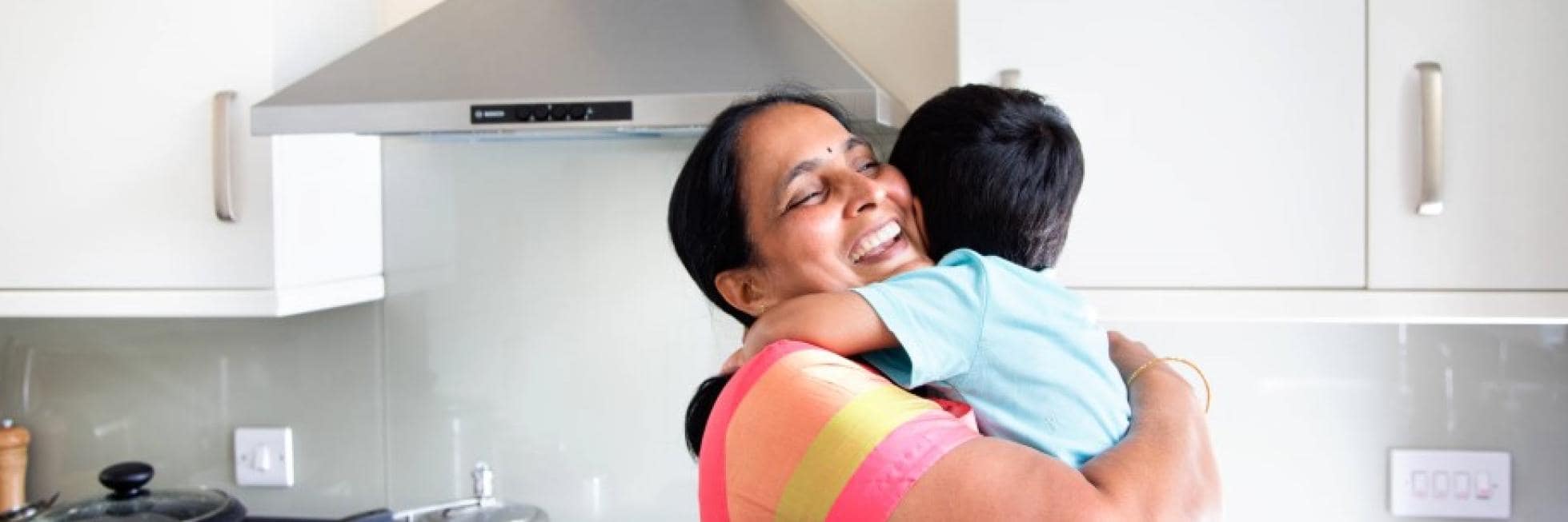 A woman happily embraces a child in a modern kitchen, with a stovetop and range hood in the background.