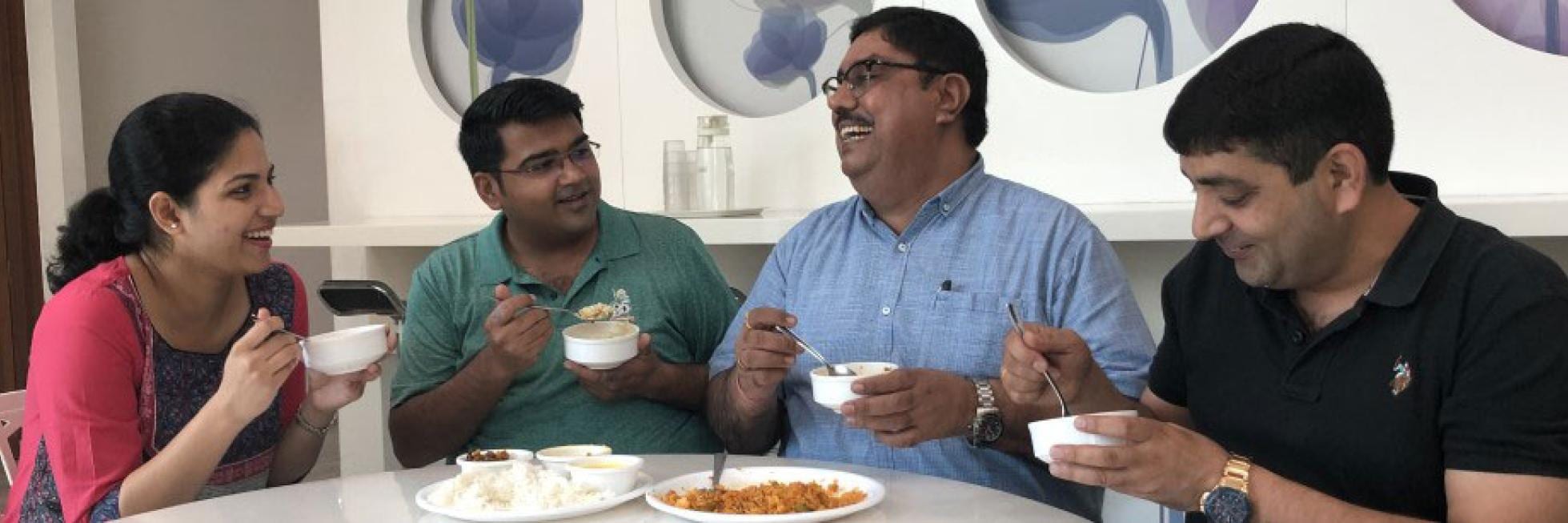 Four people are sitting at a table, sharing a meal and smiling. Plates of rice and other dishes are in front of them.