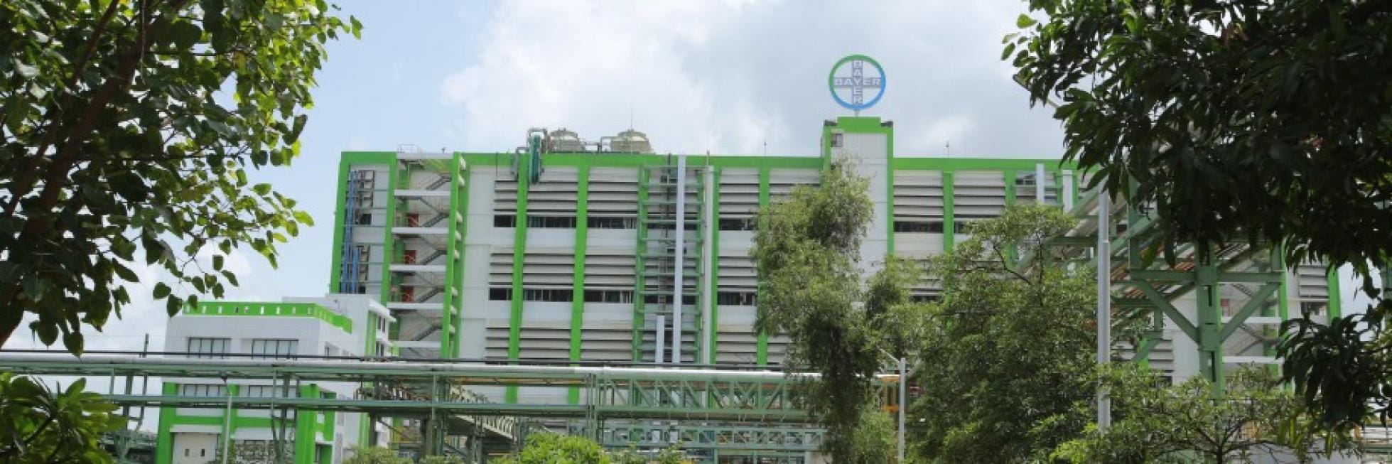 A large industrial building with a green and white facade, surrounded by trees, under a partly cloudy sky. A round sign with text is on top of the building.