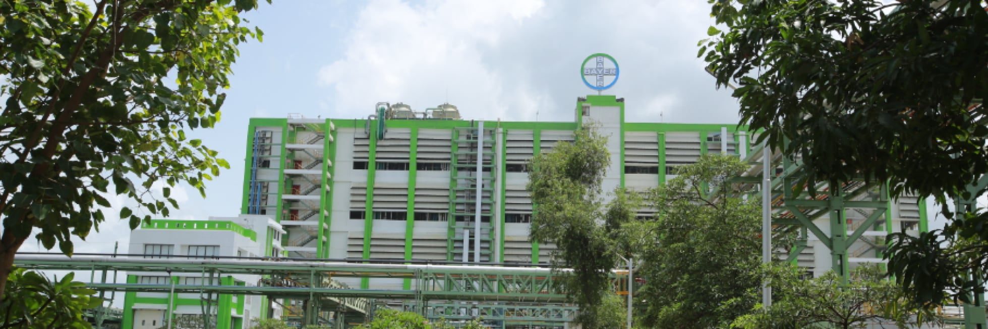 Large industrial building with green accents and a logo on top, surrounded by trees under a partly cloudy sky.