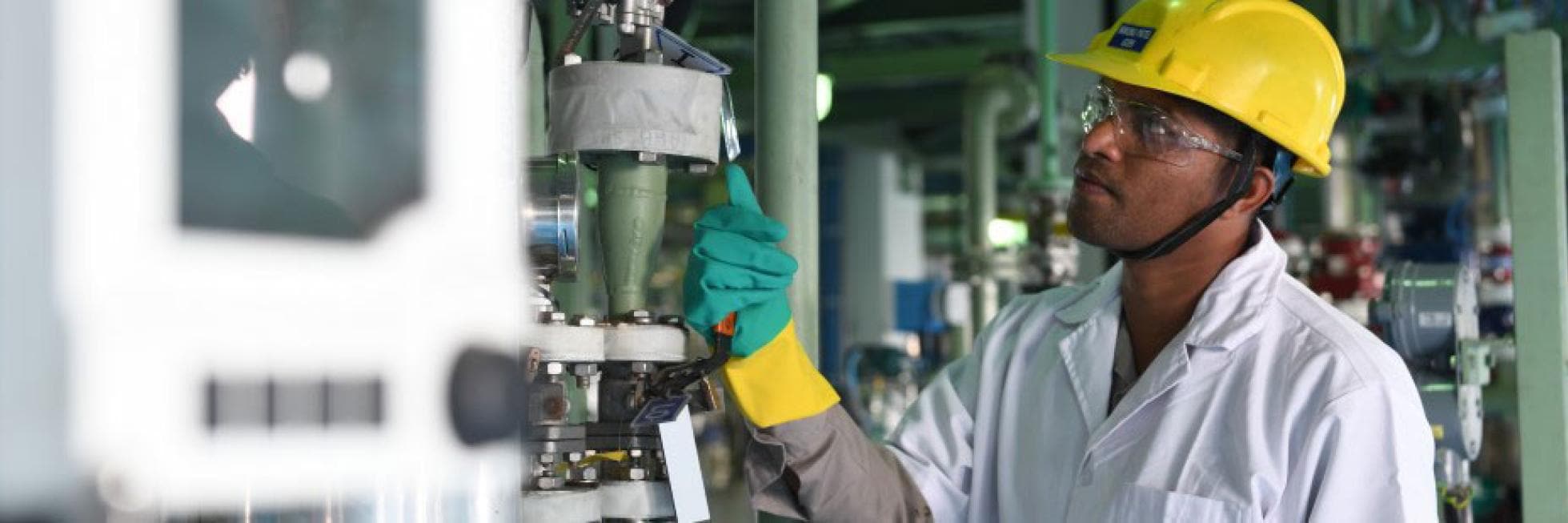 A person wearing a yellow hard hat and safety goggles inspects industrial machinery in a factory setting.