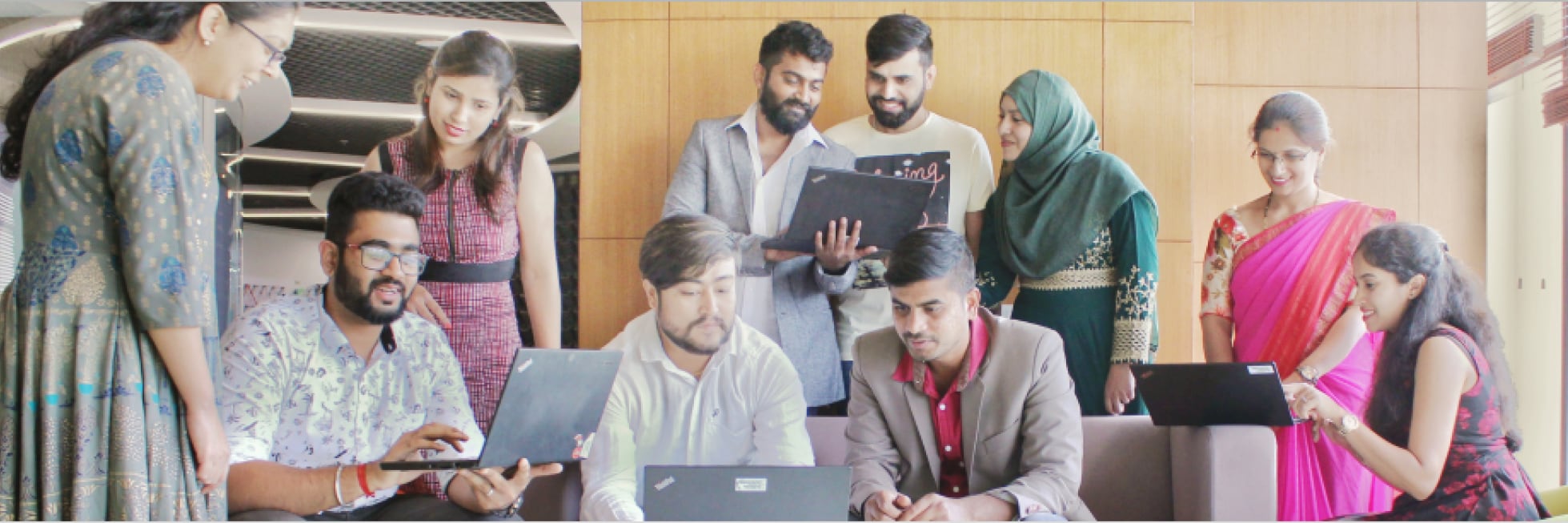 A diverse group of people collaborates with laptops, standing and sitting in a modern office setting.