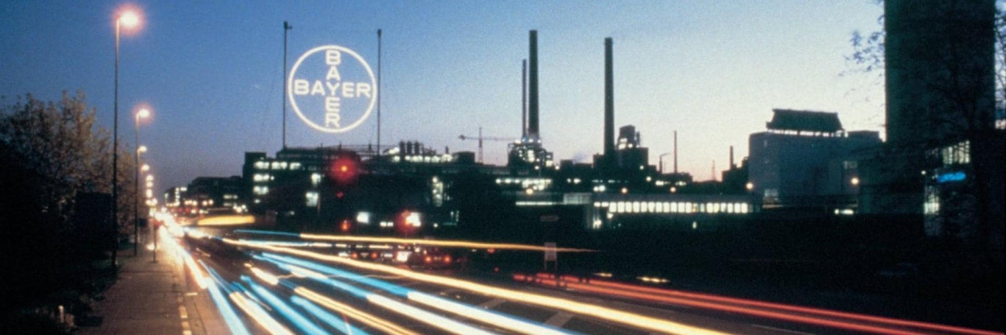 Evening view of an industrial complex with smokestacks and bright light trails from passing vehicles. The Bayer logo is prominently displayed on a large circular sign.