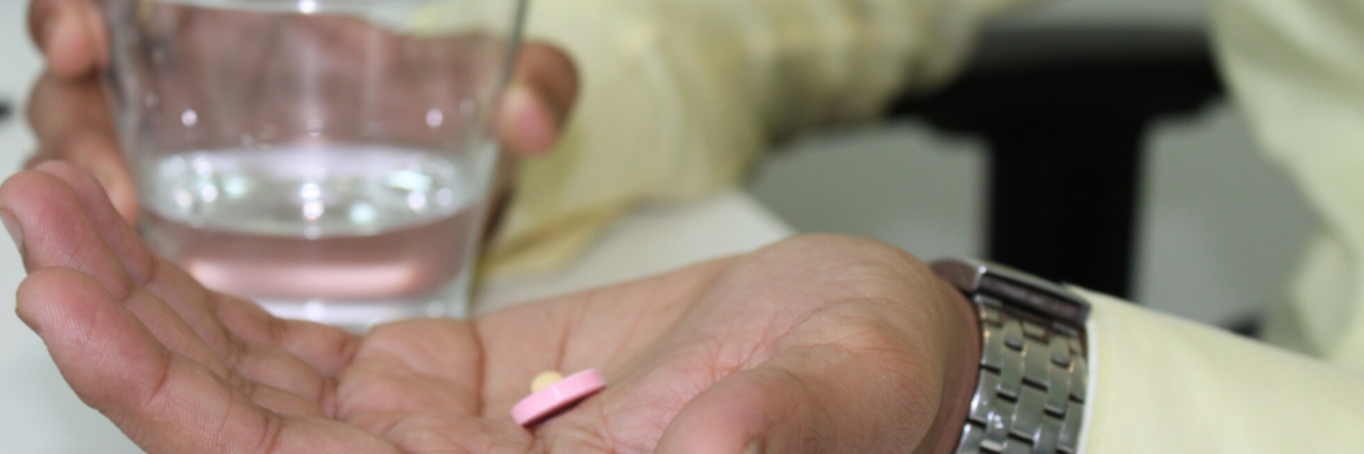 A hand holds pink and yellow pills with a glass of water in the background.