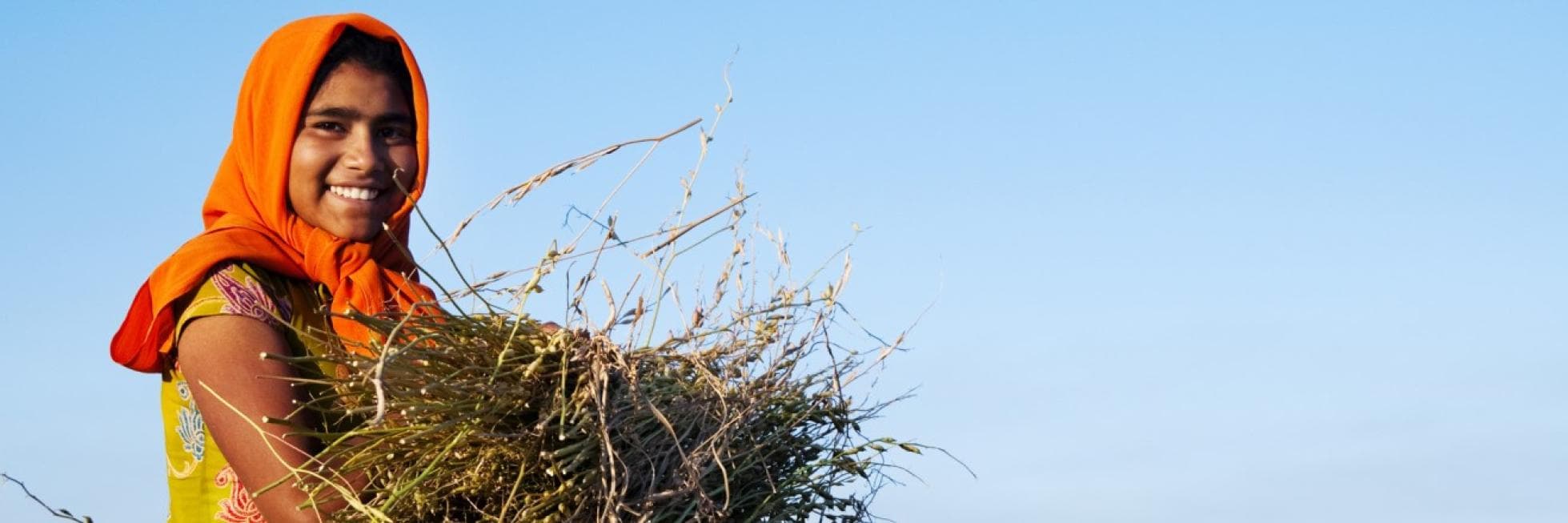 A person wearing an orange headscarf, smiling and holding a bundle of dry plants against a clear blue sky.