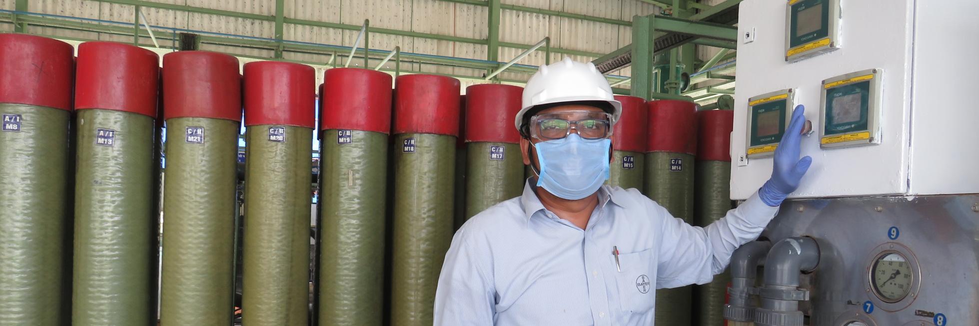 A person wearing a hard hat, safety glasses, mask, and gloves stands next to industrial equipment with cylinders in the background.