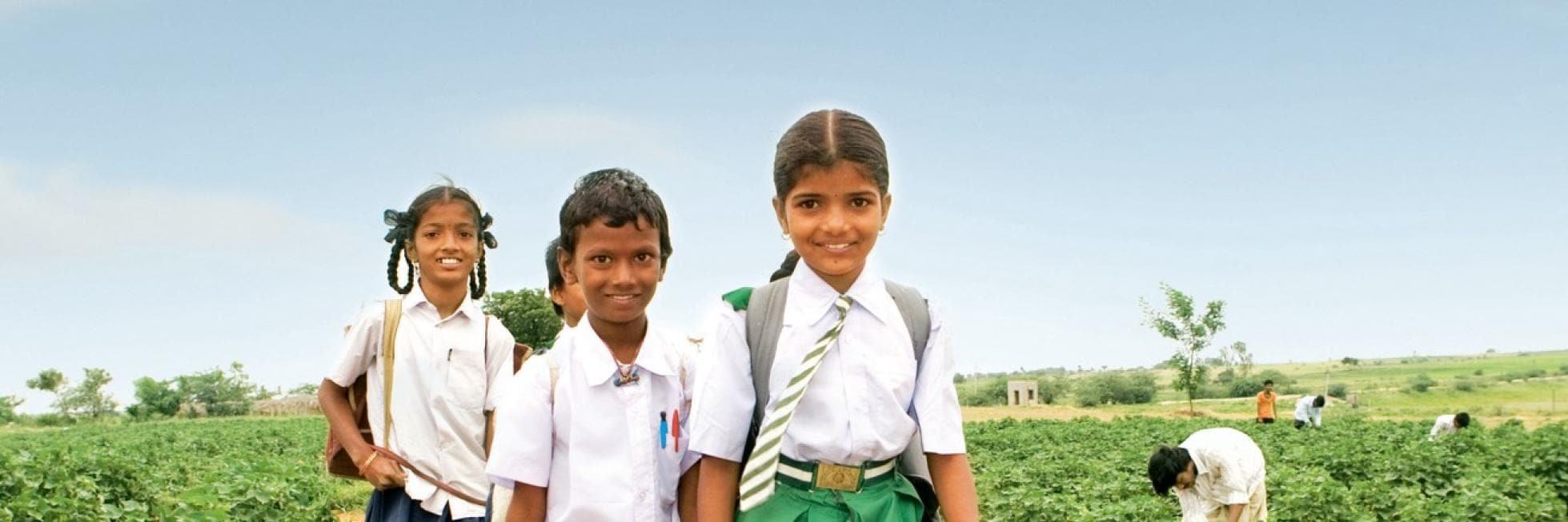 Three children in school uniforms smile in a field, with others working in the background under a clear sky.