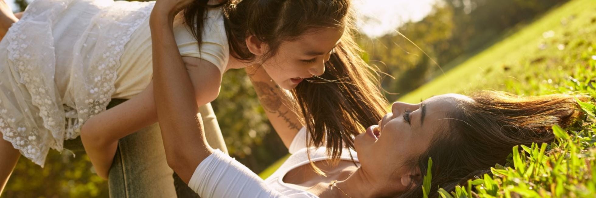 Woman lying on grass playfully lifting a smiling child in a park setting.