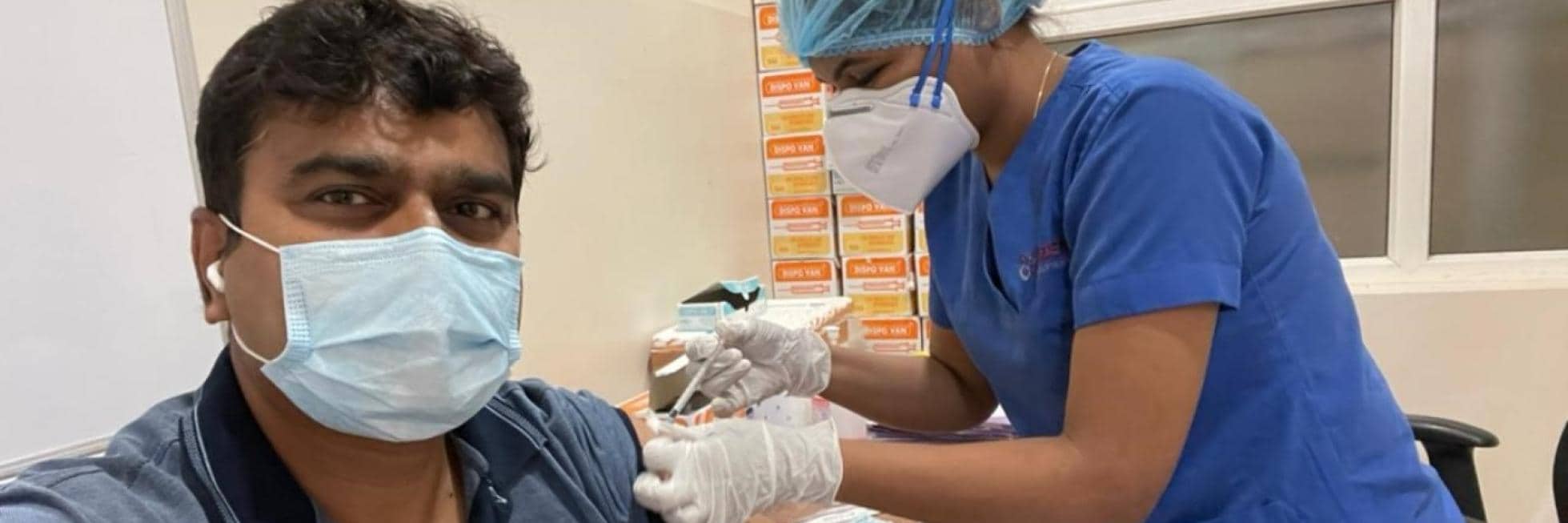 A healthcare worker in blue scrubs administers a vaccine to a seated person wearing a mask in a medical setting.