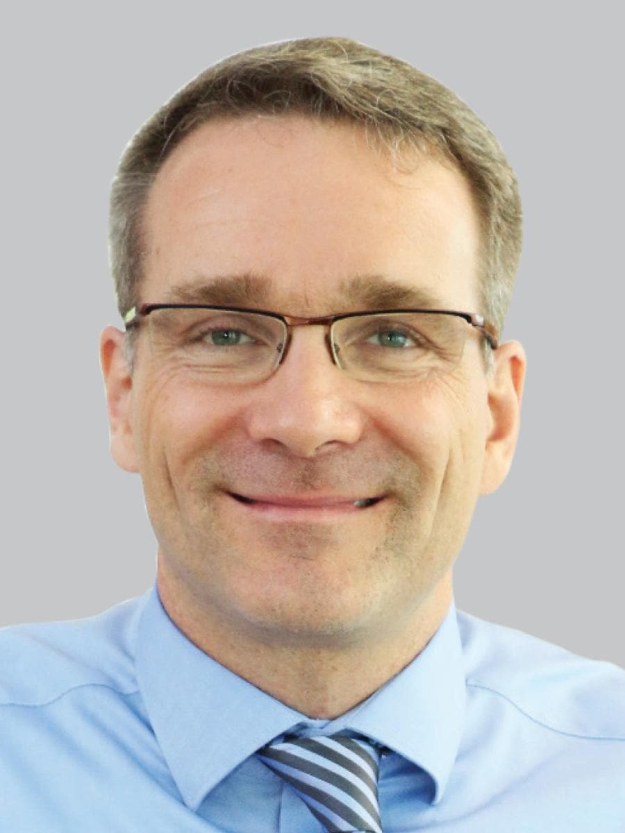 Man with glasses smiling, wearing a blue shirt and striped tie, against a plain background.
