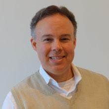 A person smiling, wearing a white shirt and beige vest, against a plain background.